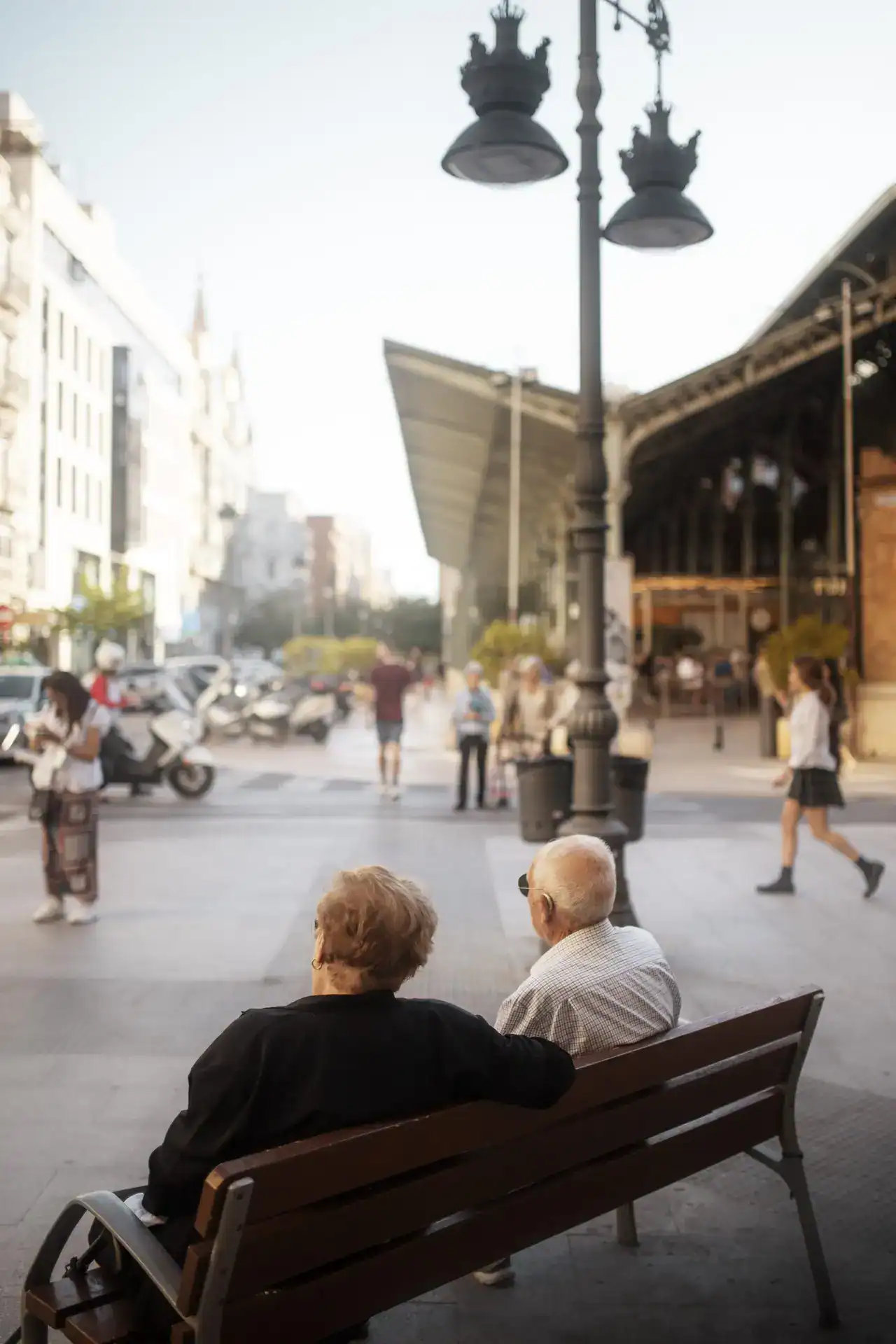תמונה של זוג קשישים יושבים על ספסל ברחוב הומה בבוקר שמש, הסביבה עירונית עם קהל הולכי רגל ורכבים על הכביש, מבנה מודרני ומנורה עם עיטורים קלאסיים, אווירה של שגרה בעיר, חיי קהילה ואווירה חמה ומשוחררת, מתאים לקידום תיירות, חיים עירוניים וקהילה.